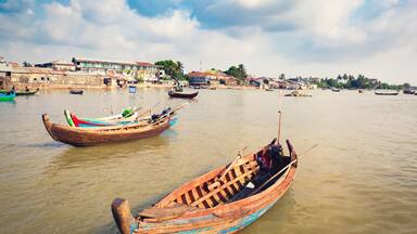 A river with traditional wooden boats, a distant shoreline with buildings, and a partly cloudy sky.