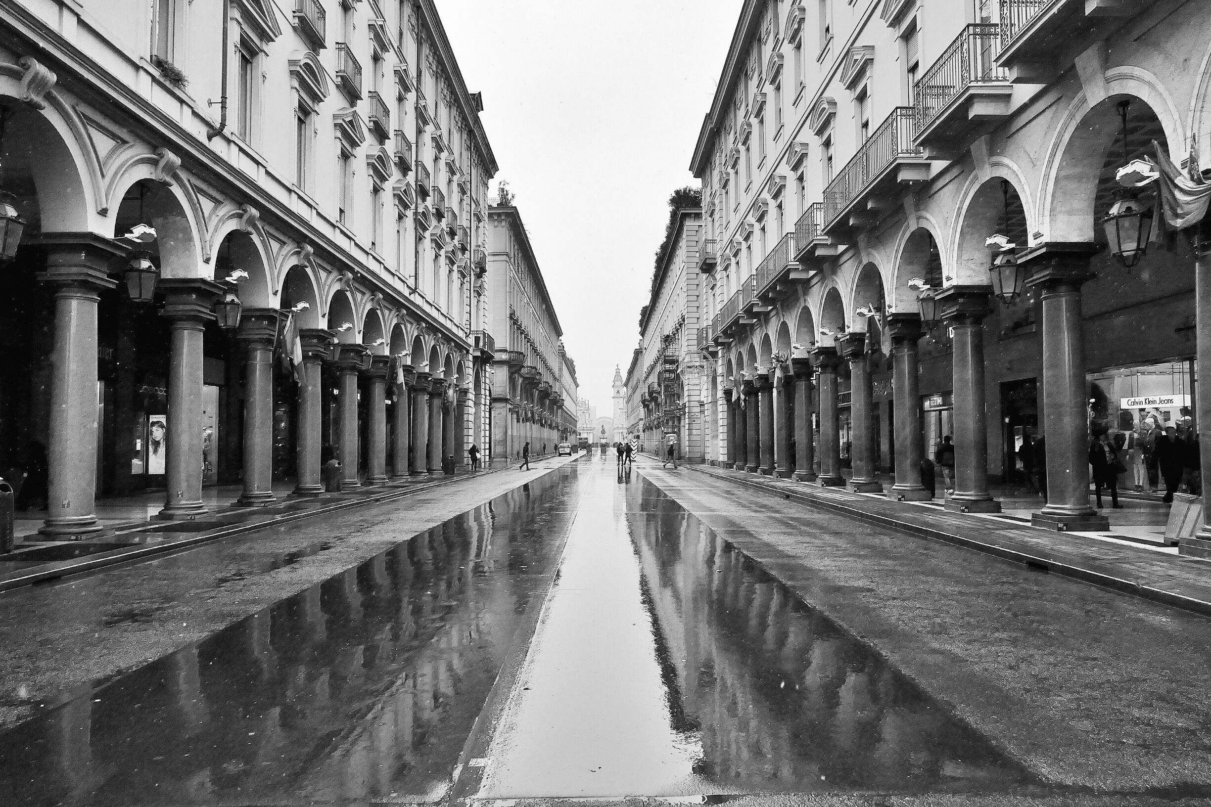 Rua molhada com pavimento refletivo, ladeada por edifícios com janelas e sacadas em arco.