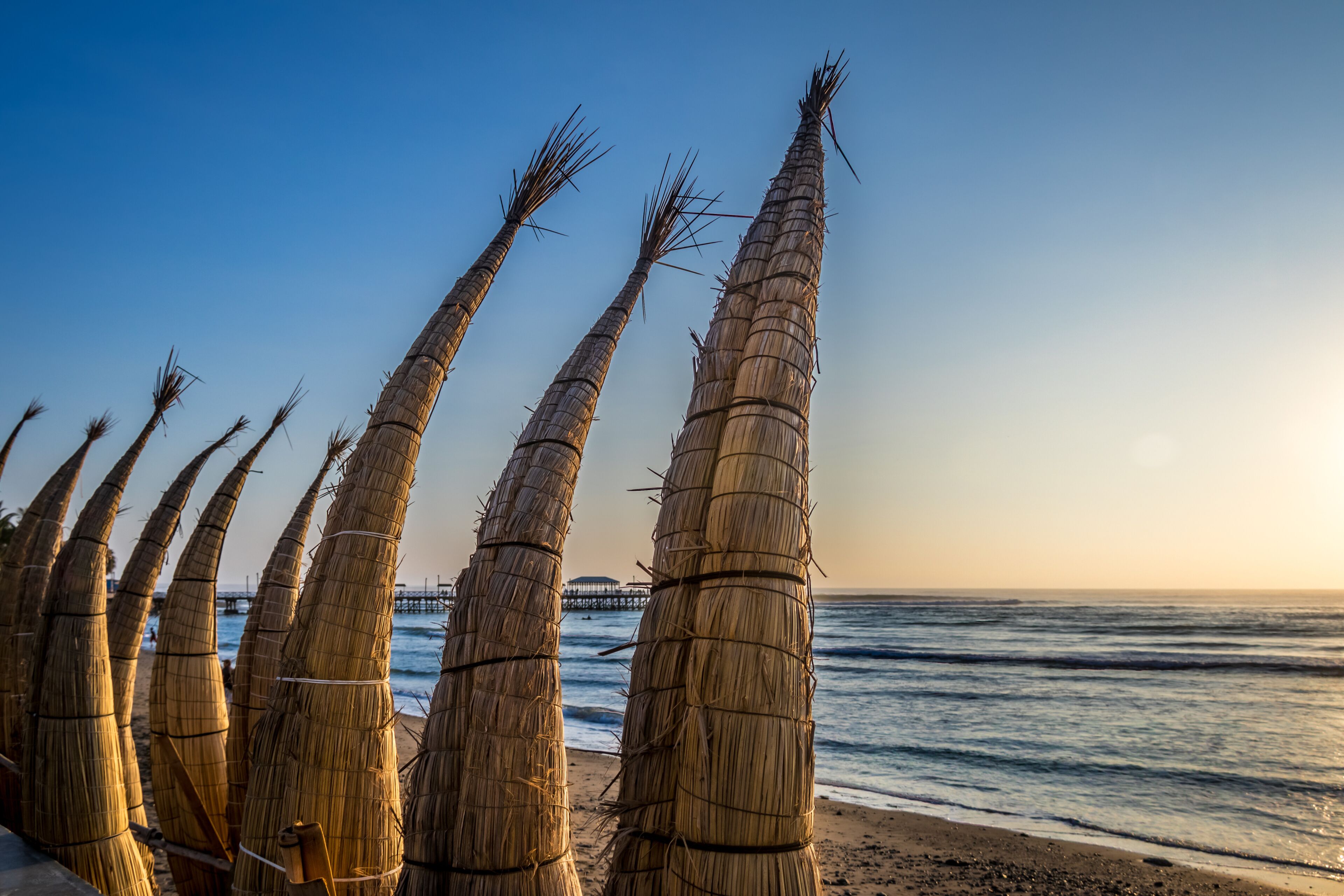 A beach with tall, bundled palm fronds and a pier in the background.