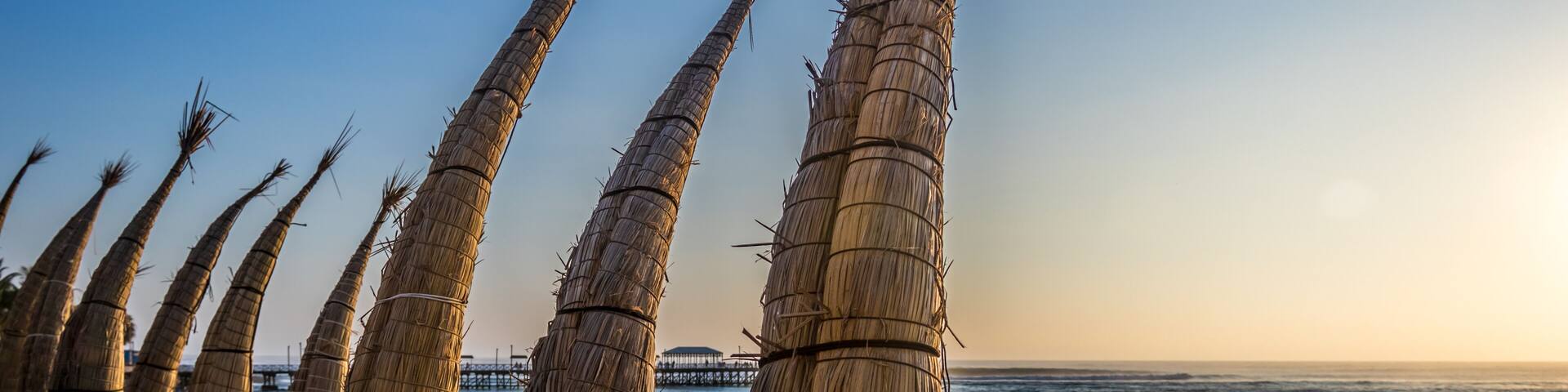 A beach with tall, bundled palm fronds and a pier in the background.
