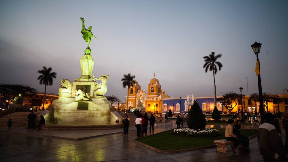 A nighttime city square with a prominent statue, palm trees, and a historic building with domes.