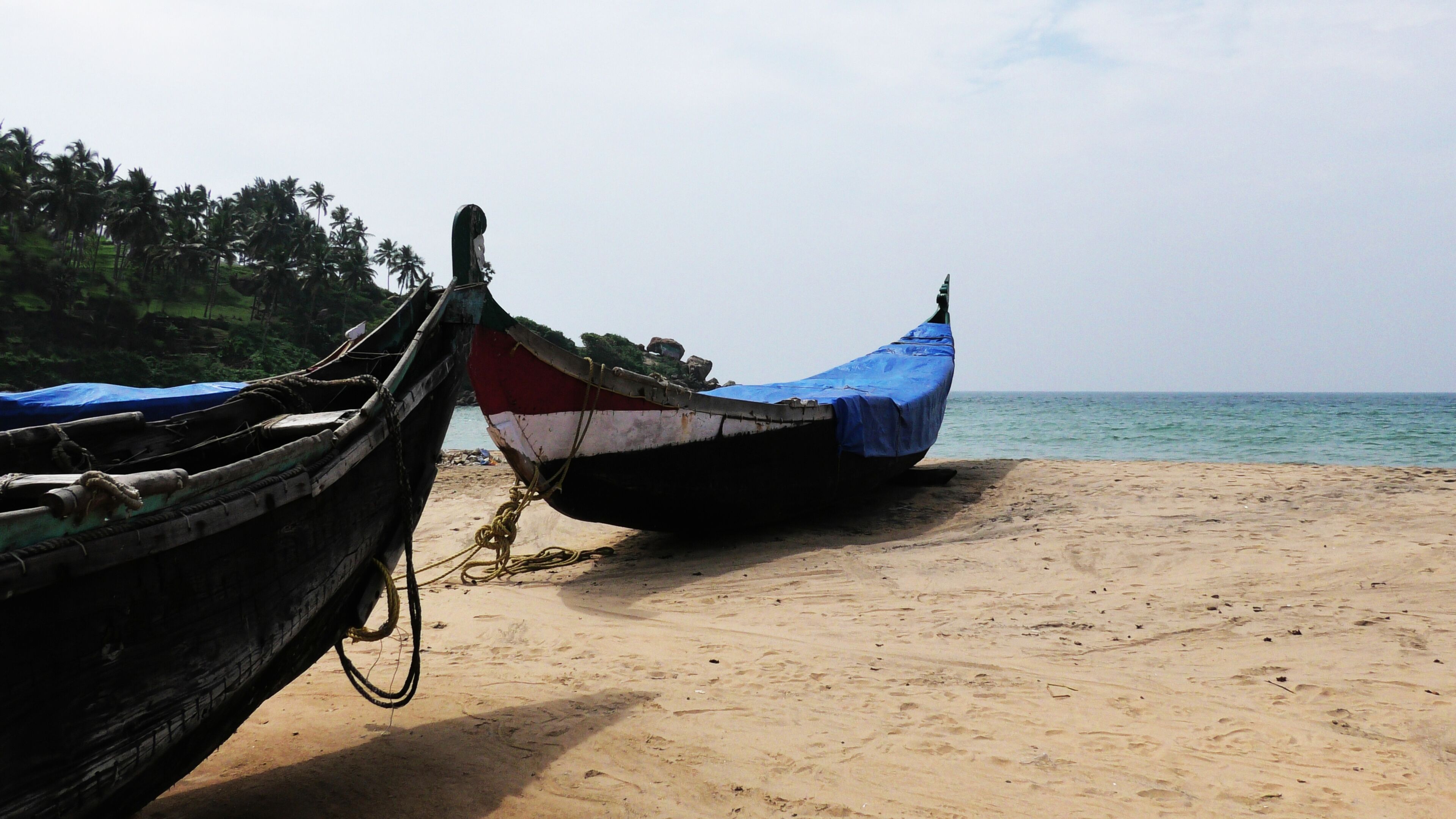 Two traditional boats tied to a sandy beach with a calm sea and palm trees in the background.