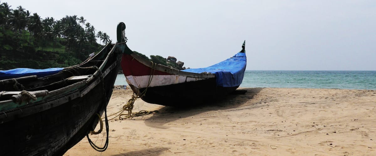 Two traditional boats tied to a sandy beach with a calm sea and palm trees in the background.