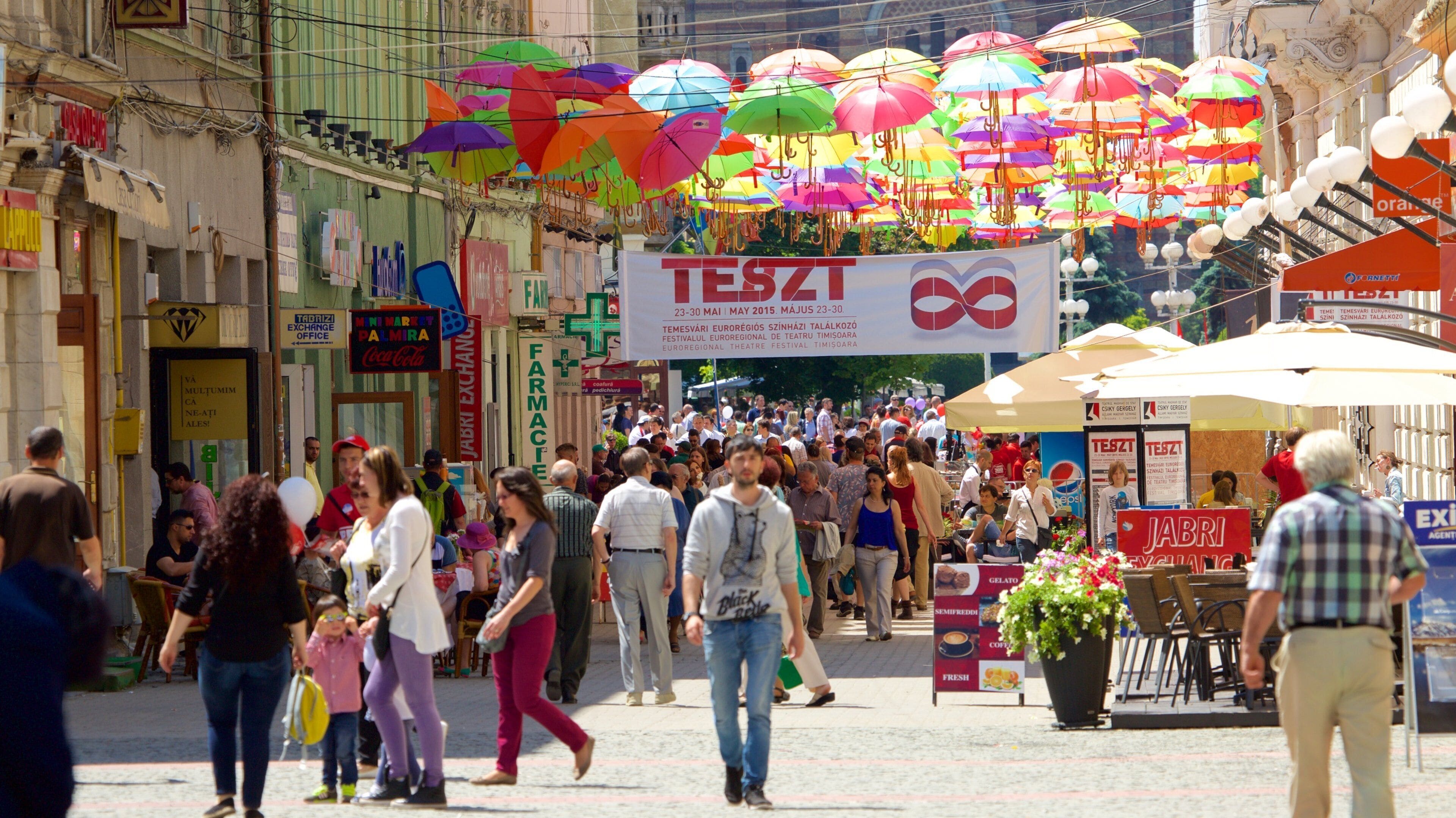 A busy street with colorful umbrellas, a crowd of people, and a sign for the 'TESZT' festival.