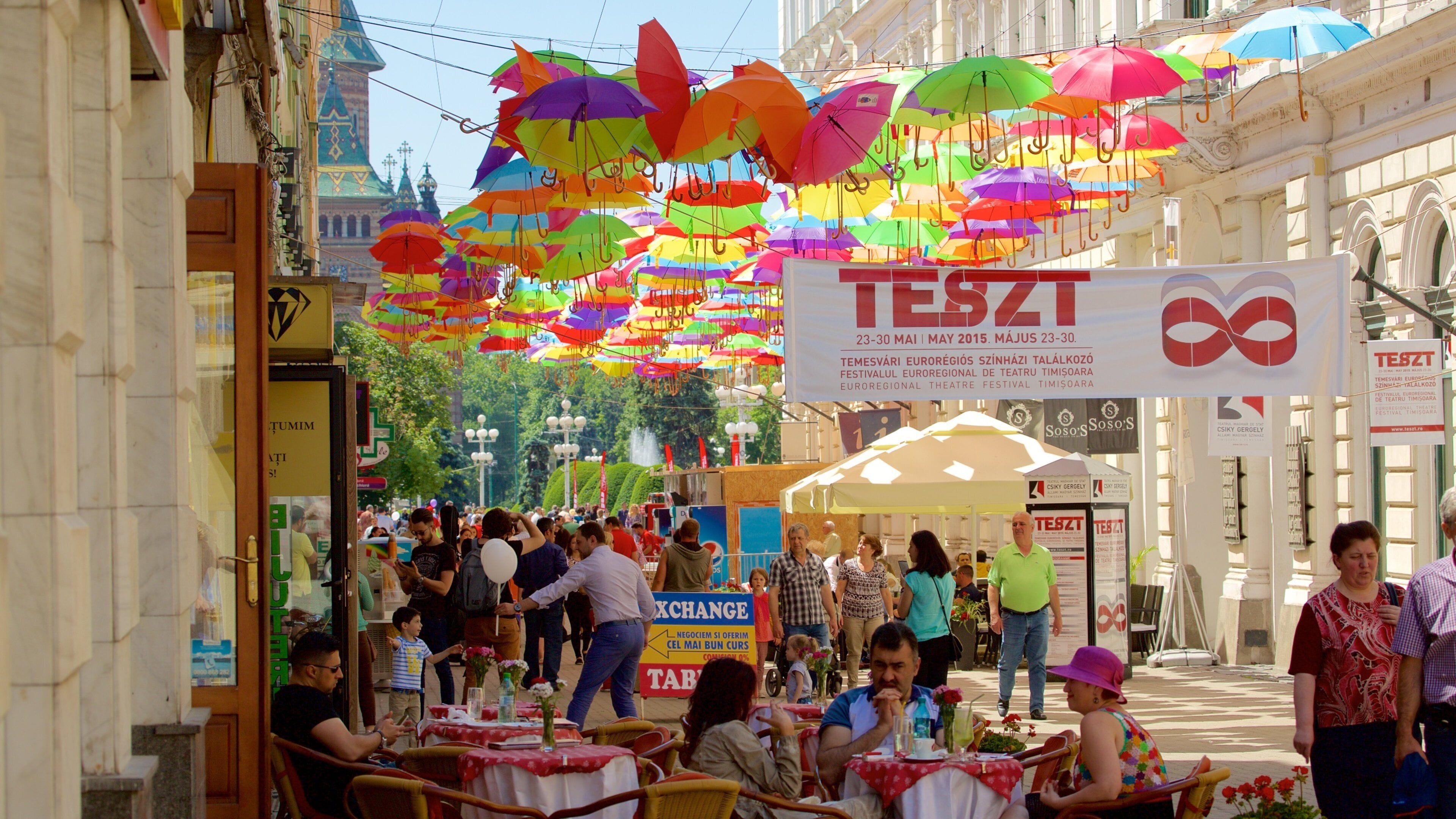 A busy street with colorful umbrellas, a sign for TESZT, and people dining at outdoor tables.