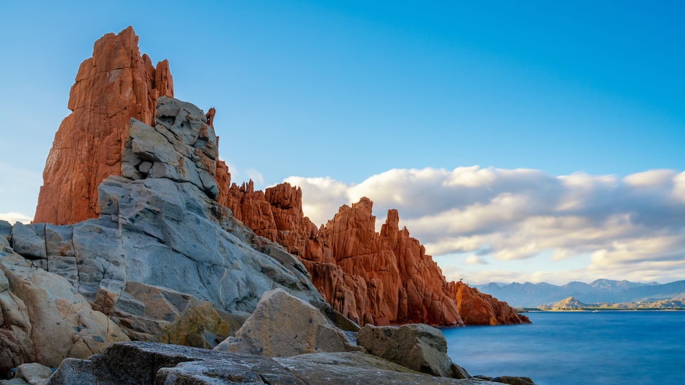 A coastal landscape with prominent red rock formations and a calm sea.