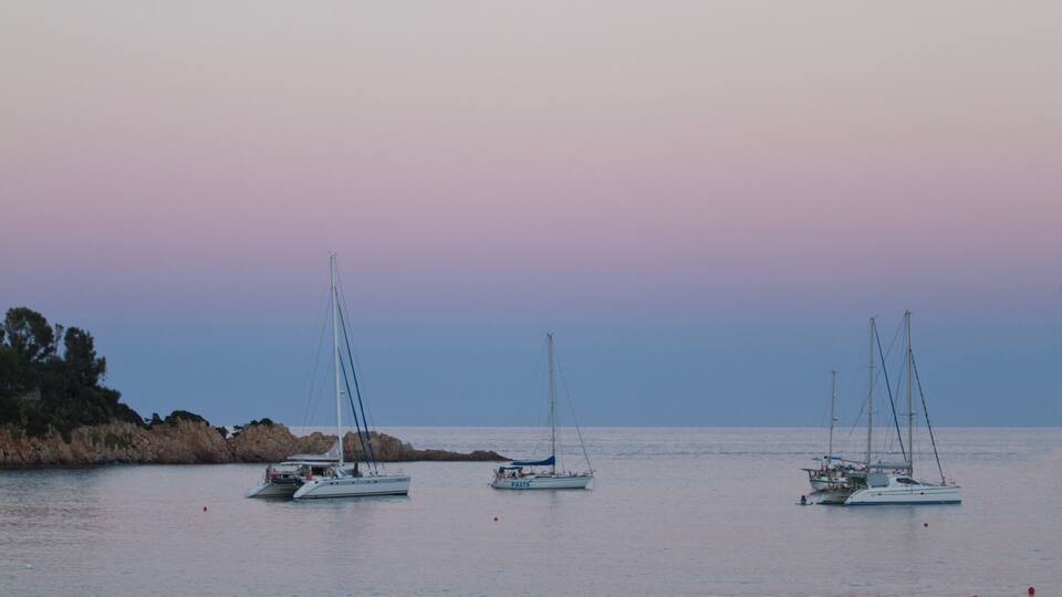 Three sailboats anchored near rocky coastline at sunset.