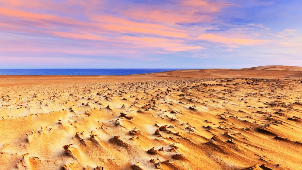 A vast desert landscape with rippled sand dunes under a colorful sky.