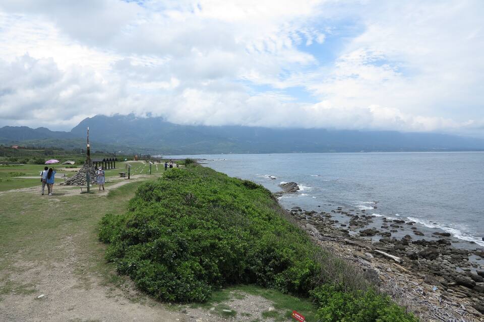 A coastal path with rocky shoreline, lush greenery, and distant mountains.