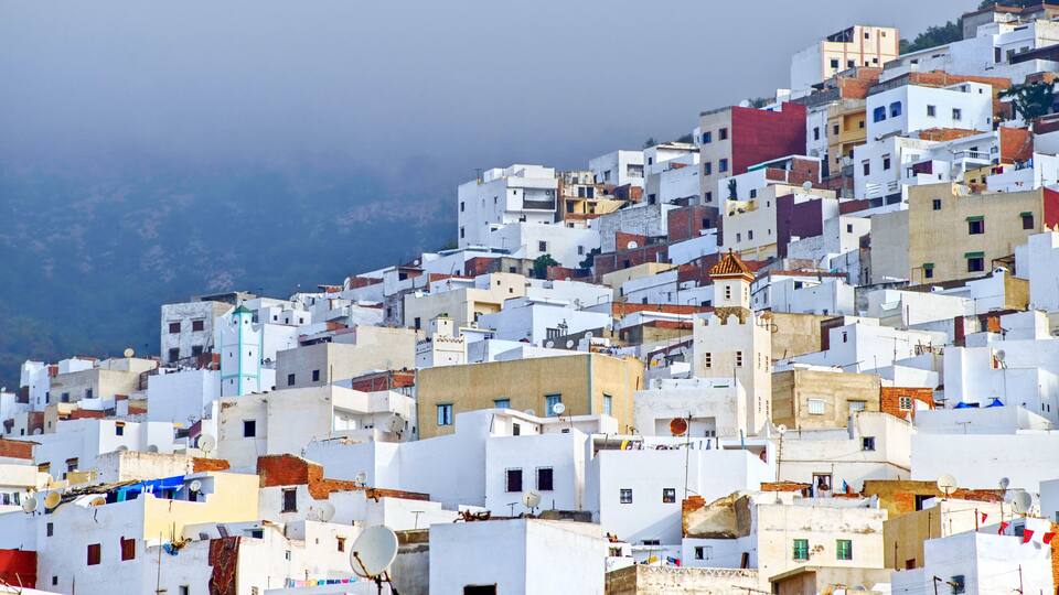 A densely packed hillside settlement with white buildings, some with red roofs, and a mountainous backdrop.