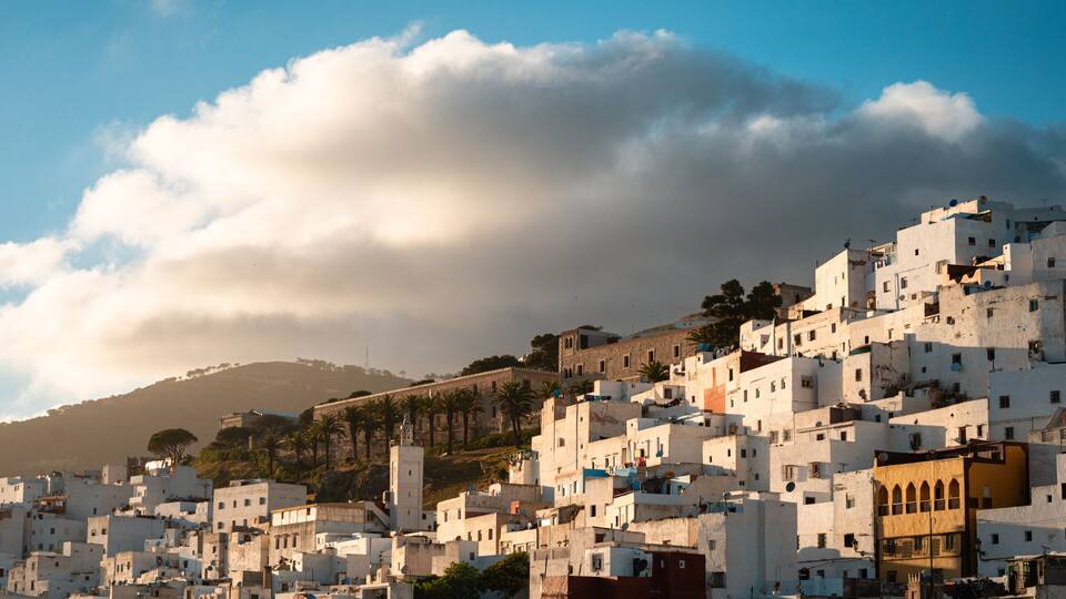 A densely packed hillside town with white buildings, a prominent tower, and a hill in the background.