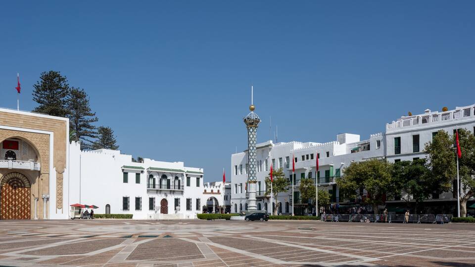 A plaza with a large ornate gate, a central tower, and white buildings with red flags.