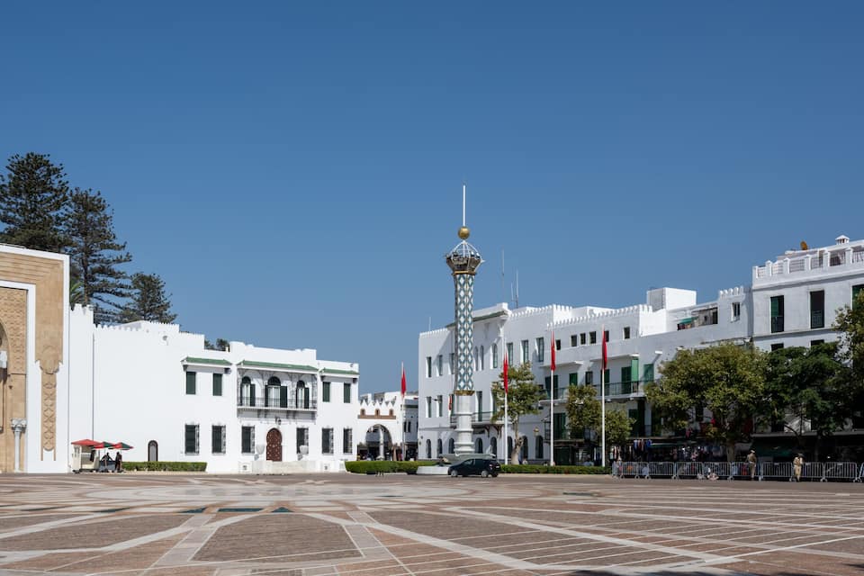 Una plaza con una imponente puerta decorada, una torre central y edificios blancos adornados con banderas rojas.