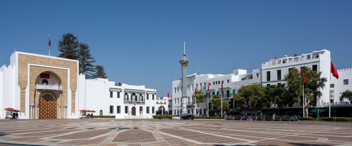 A plaza with a large ornate gate, a central tower, and white buildings with red flags.