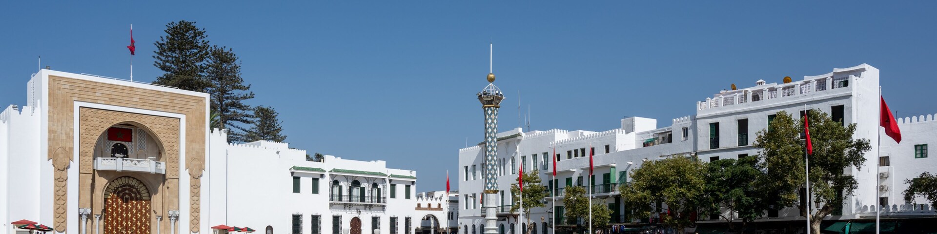 A plaza with a large ornate gate, a central tower, and white buildings with red flags.