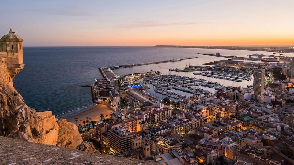 A coastal cityscape at sunset with a historic lighthouse, marina, and densely packed buildings.