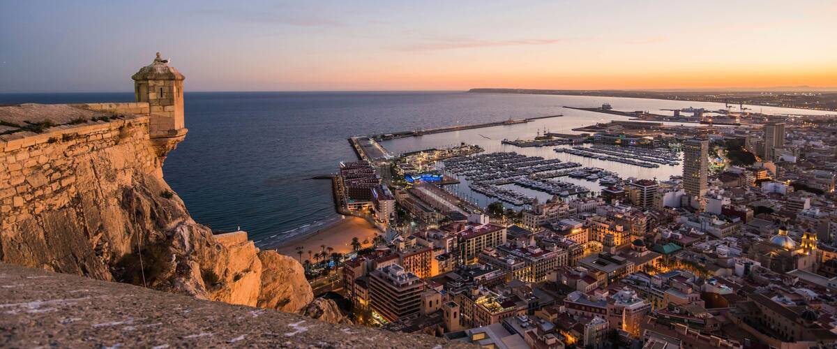 A coastal cityscape at sunset with a historic lighthouse, marina, and densely packed buildings.