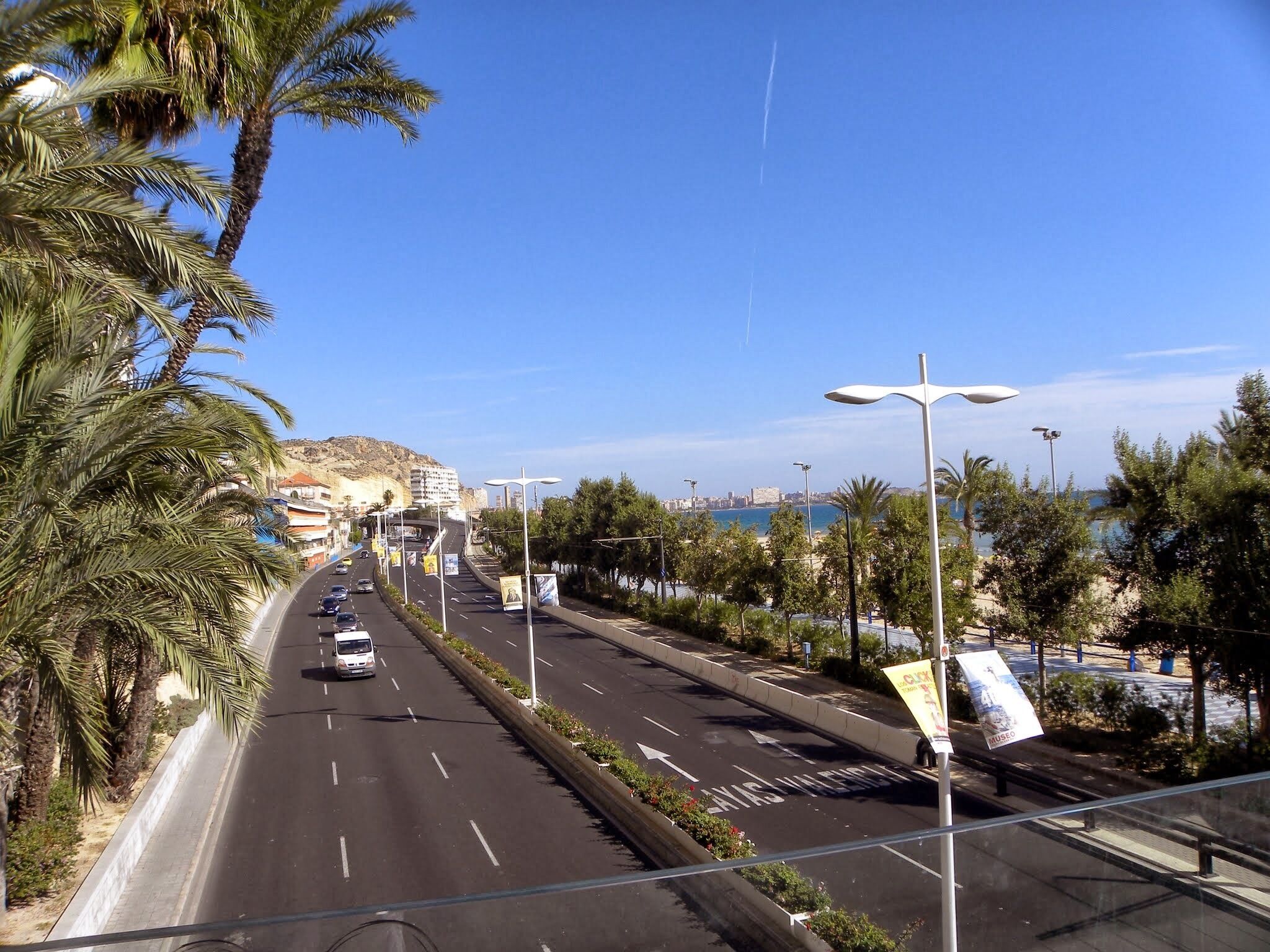 A coastal road with palm trees, a clear blue sky, and a distant view of a city and sea.
