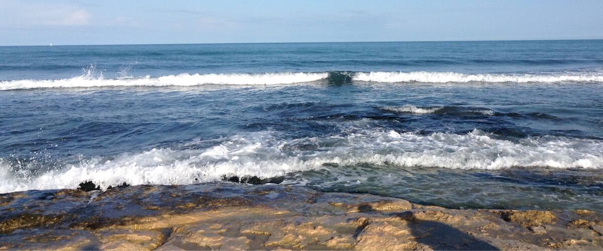 A rocky shoreline with waves crashing onto the beach under a clear blue sky.