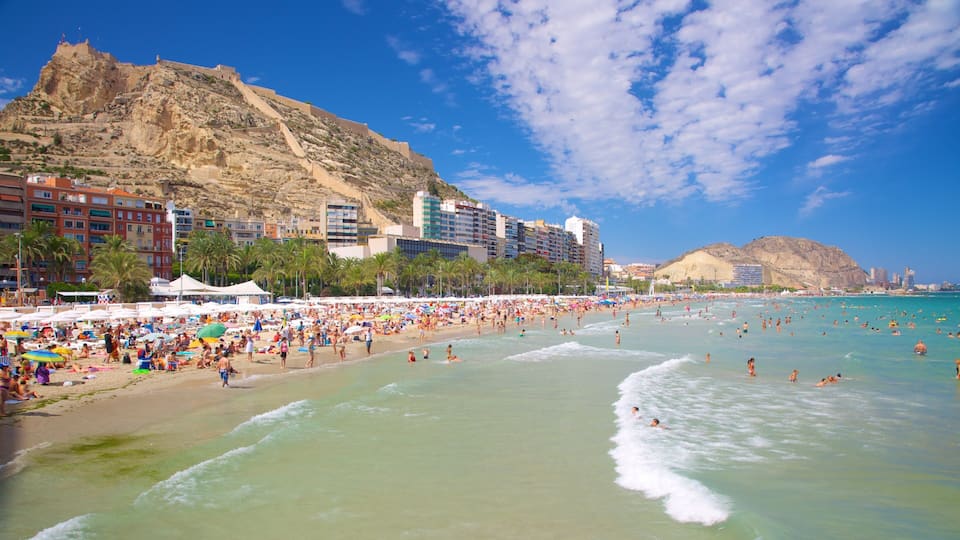 A crowded beach with people sunbathing and swimming, palm trees, and buildings in the background.