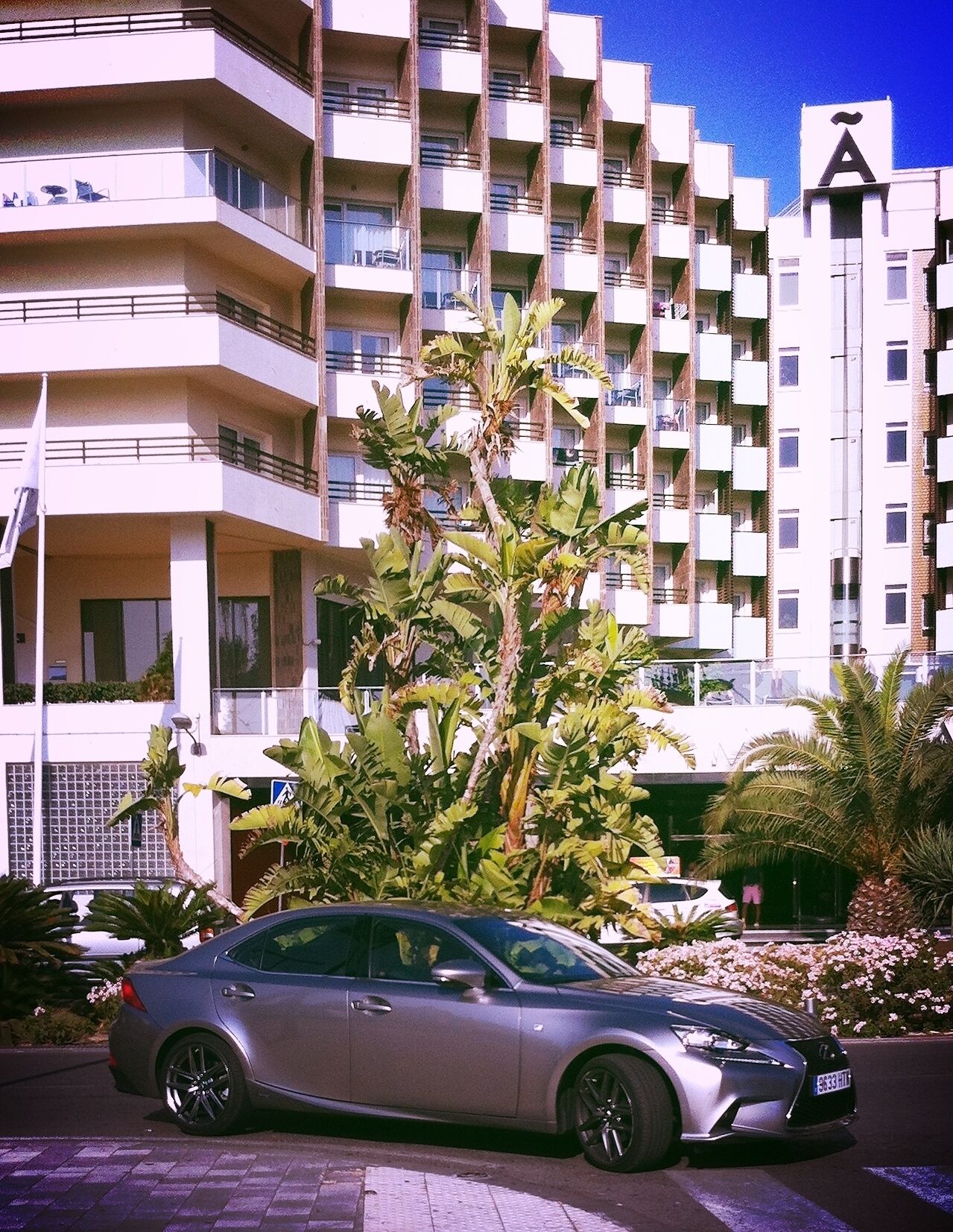 Un coche plateado estacionado frente a un edificio de varios pisos con balcones, rodeado de plantas y flores tropicales.
