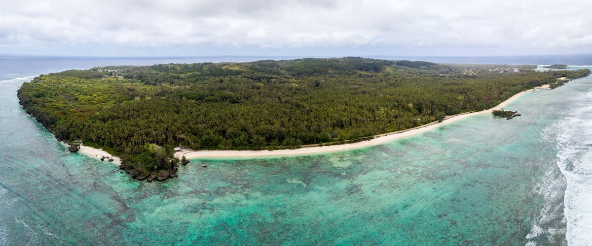 An aerial view of a tropical island with a sandy beach, clear turquoise waters, and dense green forest.