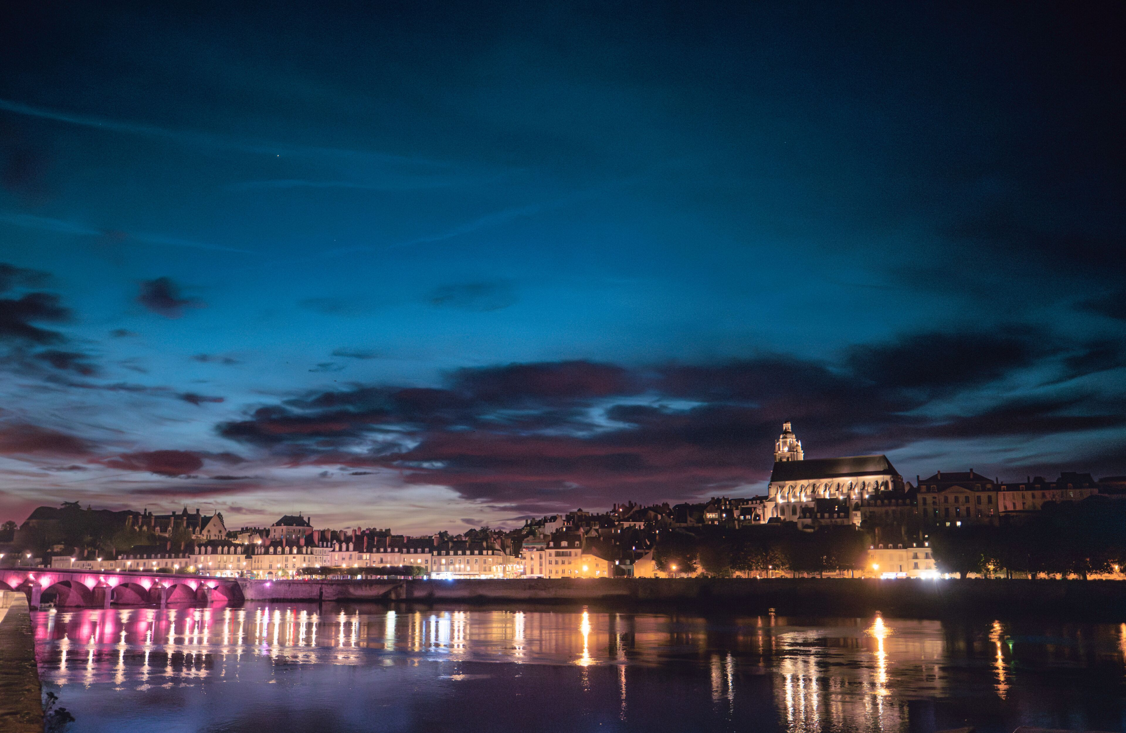 A nighttime cityscape with a prominent illuminated building, a bridge with pink lights reflecting on the water, and a river.