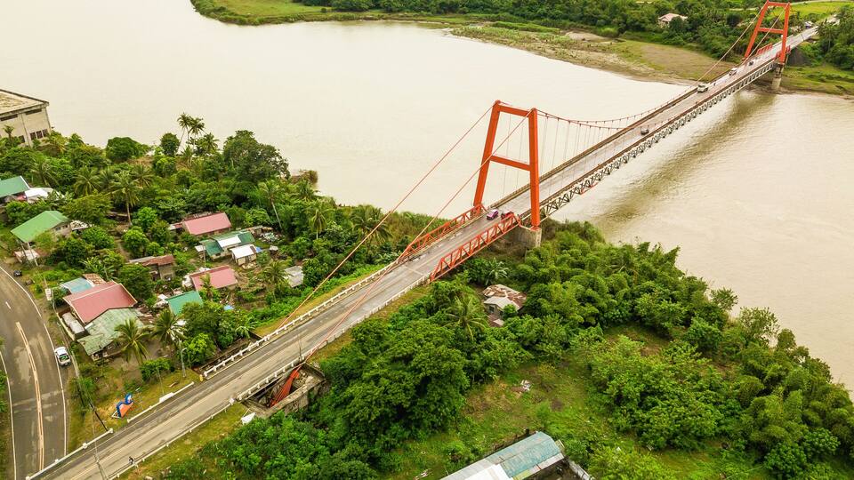 Magapit Suspension Bridge and Cagayan River, largest by volume and longest river in the Philippines. Aerial shot of wide meandering river and red bridge.