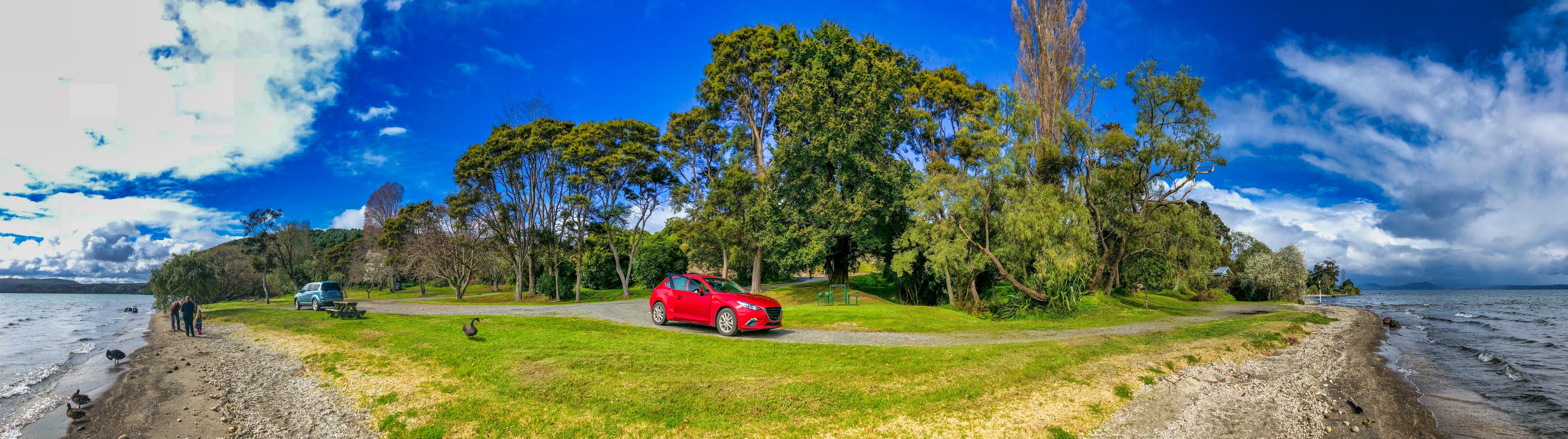 A red car parked on a grassy area near a lake with trees and a picnic table.