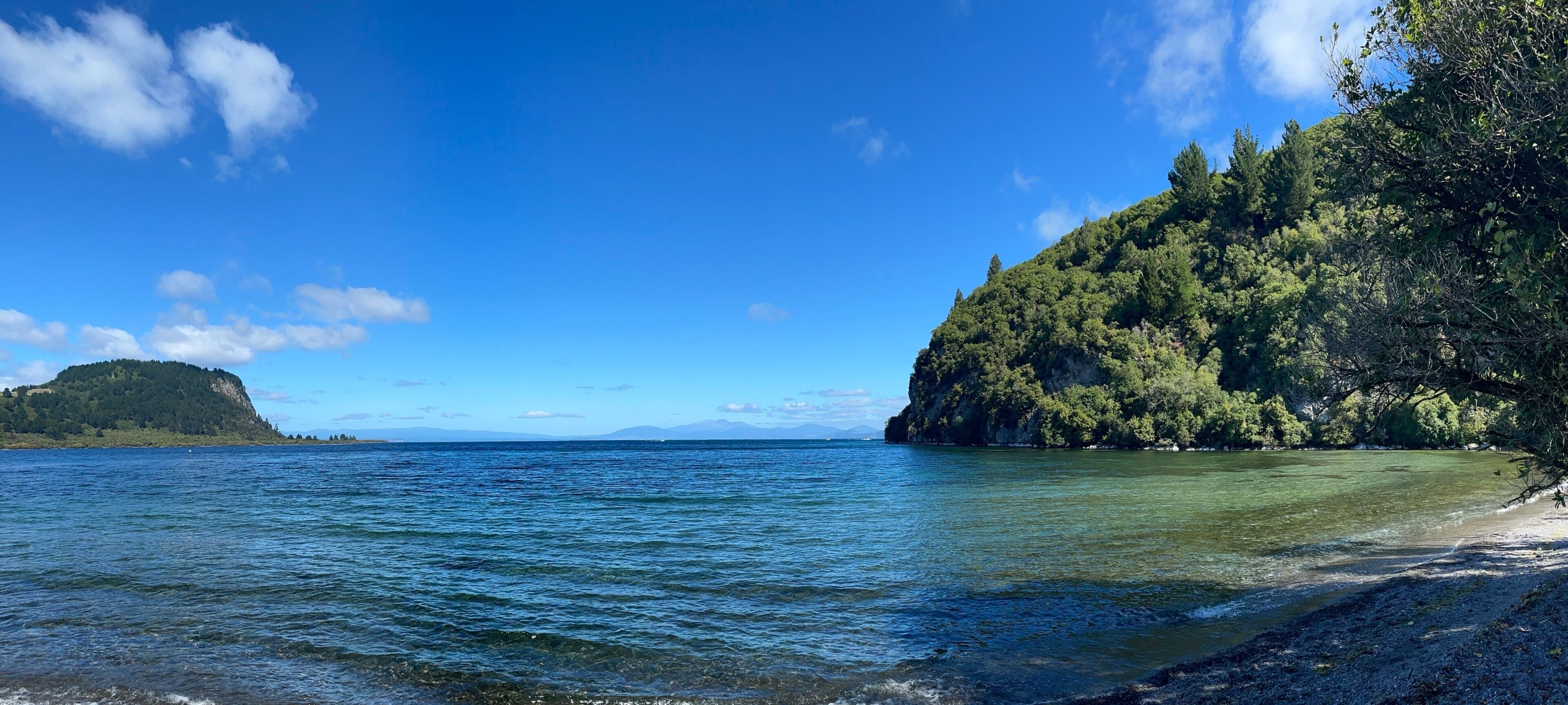 A coastal landscape with clear blue waters, a sandy beach, and a forested hill.