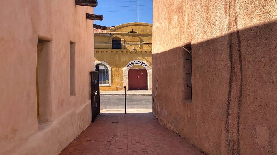 A narrow alleyway with a red brick pavement, flanked by buildings with beige walls and wooden beams. A yellow building with a red door and arched windows is visible in the distance.
