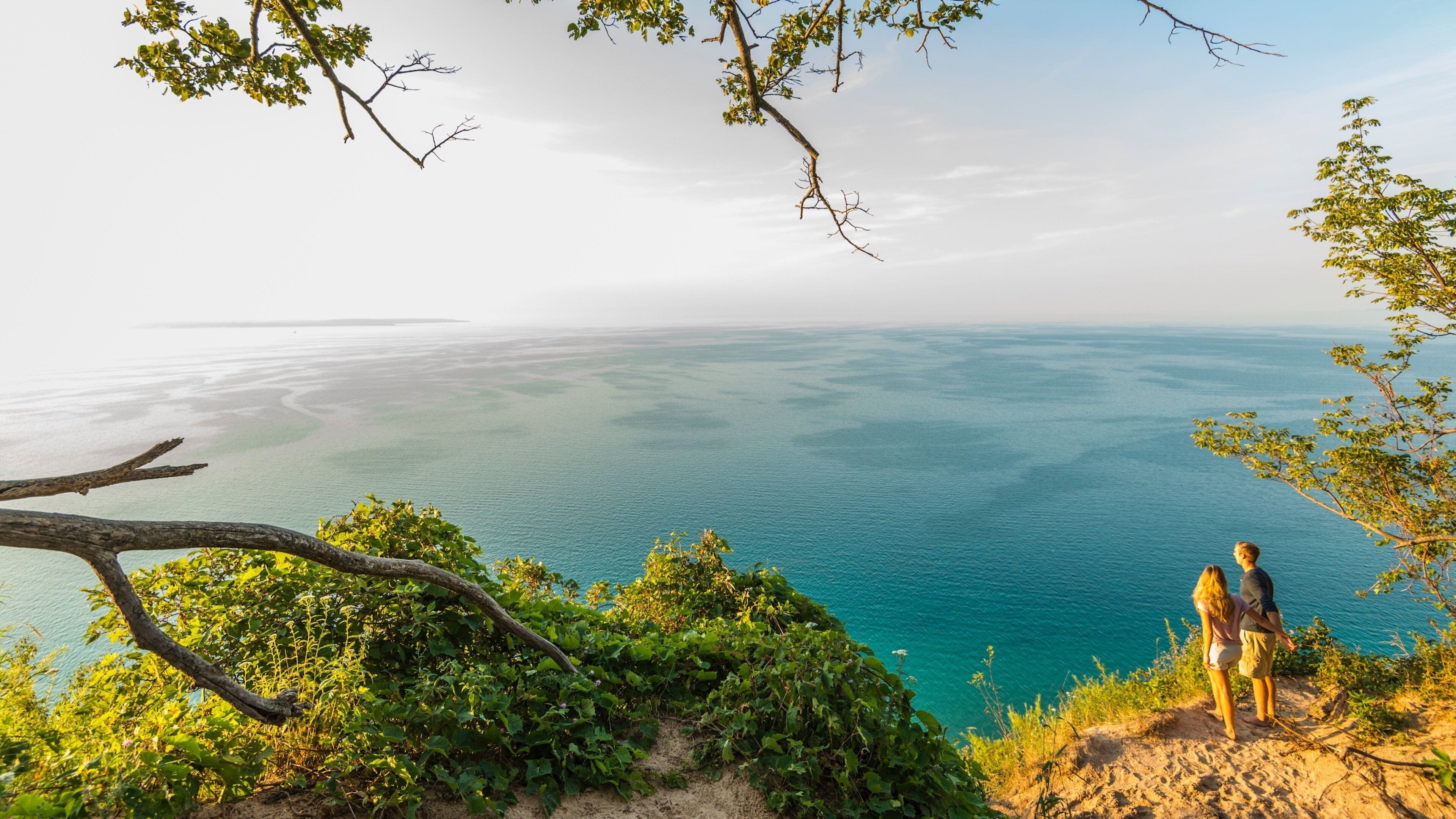 A couple overlooking a vast, clear blue sea from a sandy cliff with greenery and trees.