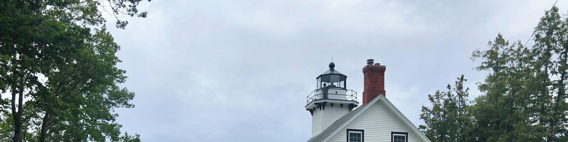 A white lighthouse with a black top, surrounded by greenery and a sign indicating it is the Mission Point Lighthouse, established in 1870.