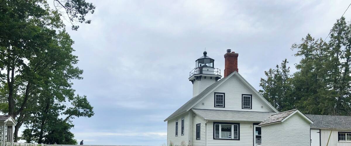 A white lighthouse with a black top, surrounded by greenery and a sign indicating it is the Mission Point Lighthouse, established in 1870.