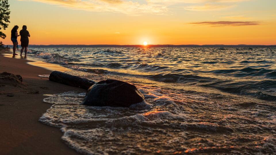 A sunset over the ocean with waves, a beach, and two people walking.
