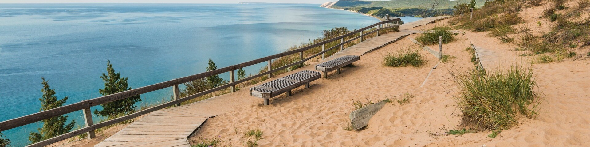 Um calçadão de madeira leva a uma praia arenosa, com bancos, oferecendo vista para um mar tranquilo e colinas ao longe.