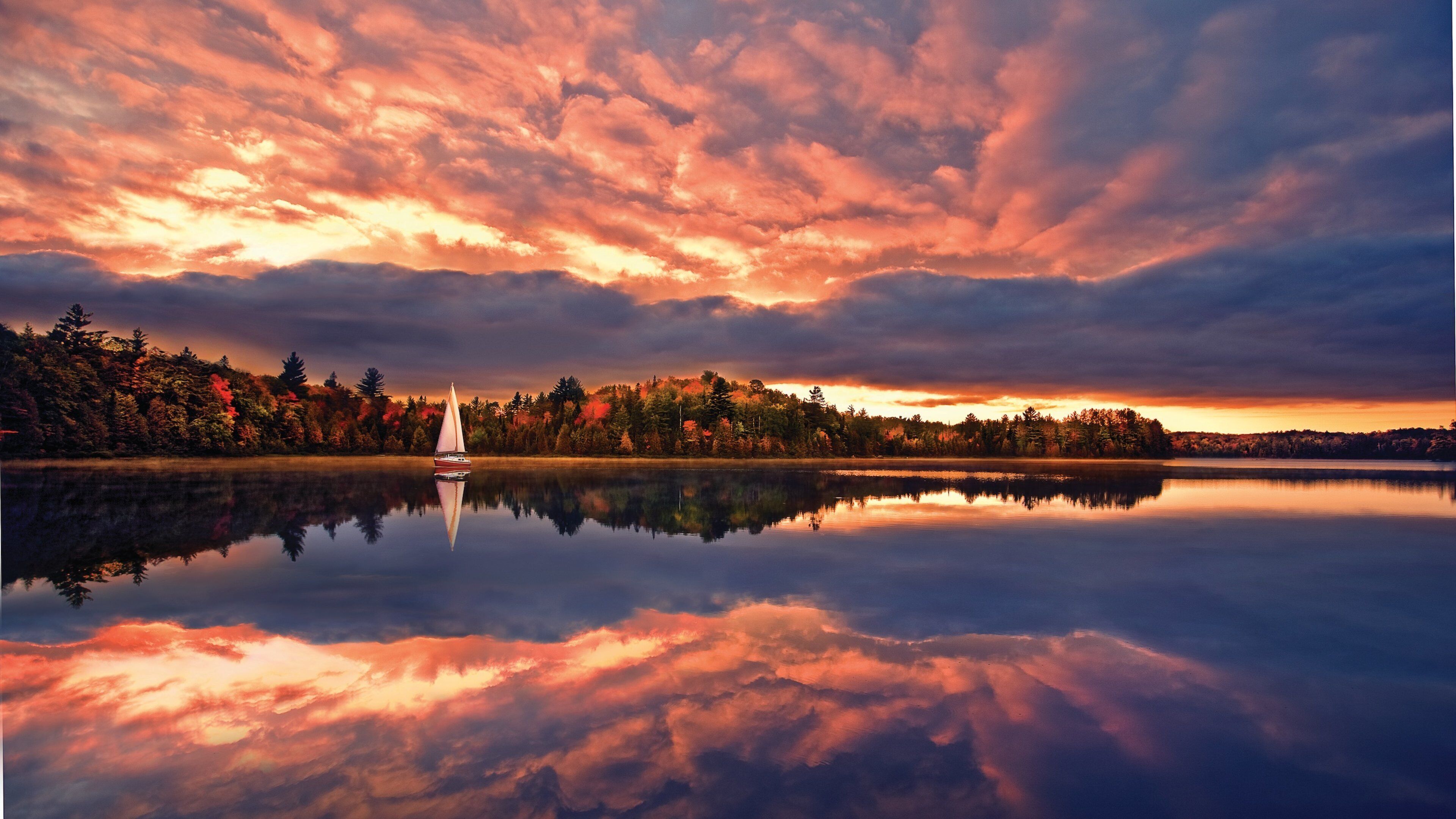 A sailboat on a calm lake with autumn-colored trees and a dramatic sky.