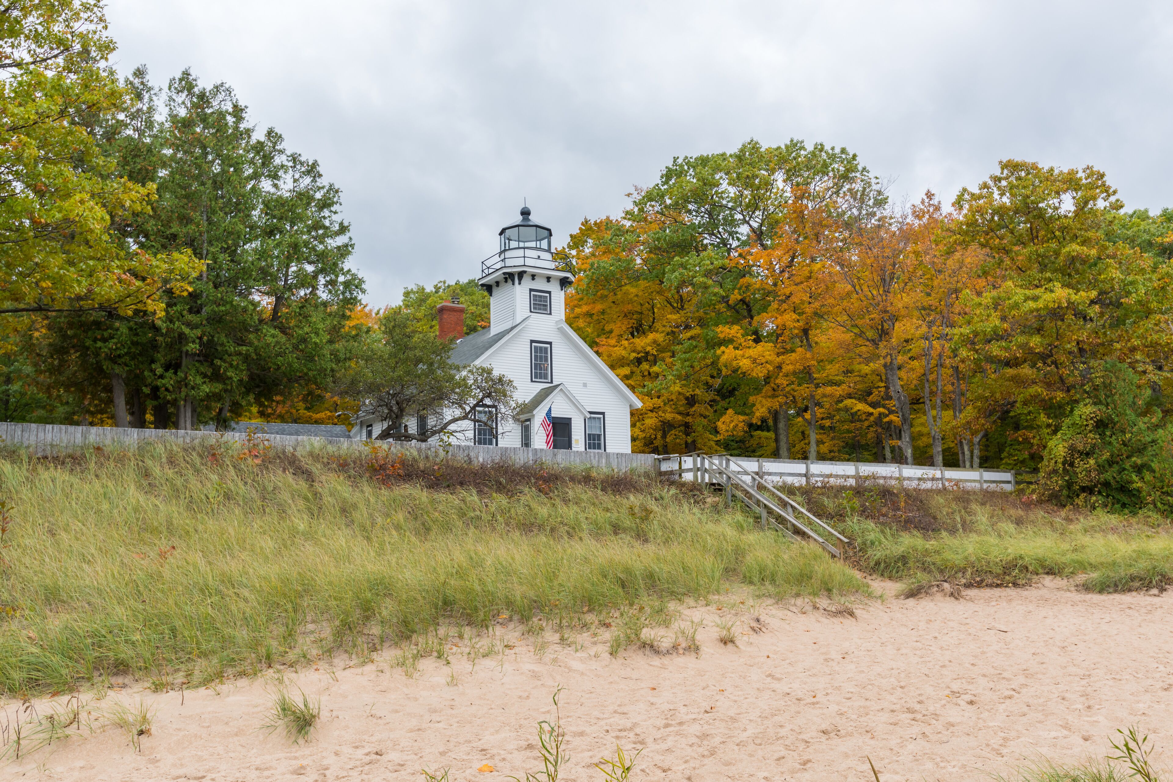 A white lighthouse with a red roof, surrounded by autumn-colored trees and a sandy beach.