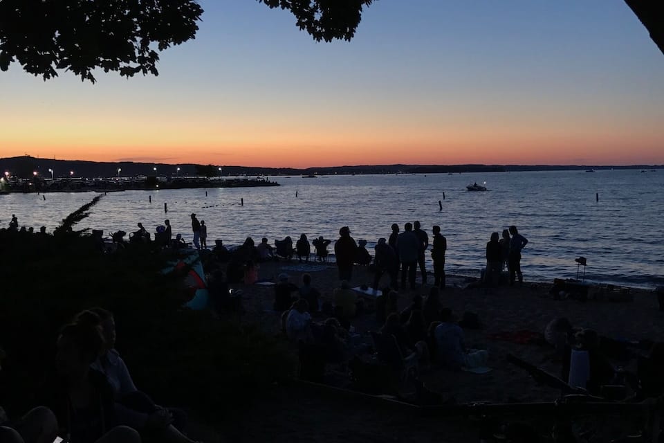 A beach at sunset with people sitting and standing near the water.