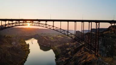 Un puente de acero arqueado sobre un río, con un atardecer de fondo.