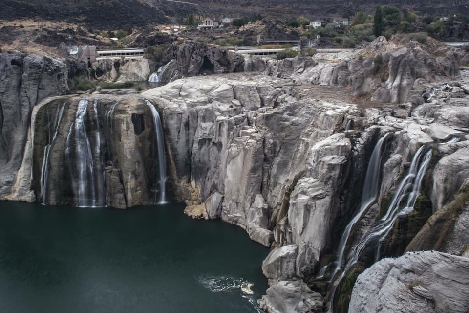 Ein großer Wasserfall stürzt über steinige Klippen in ein Gewässer, im Hintergrund sind Gebäude und Infrastruktur zu sehen.