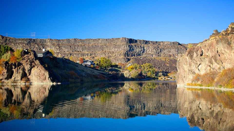 A calm lake reflecting a rocky cliff and a house with a garden.