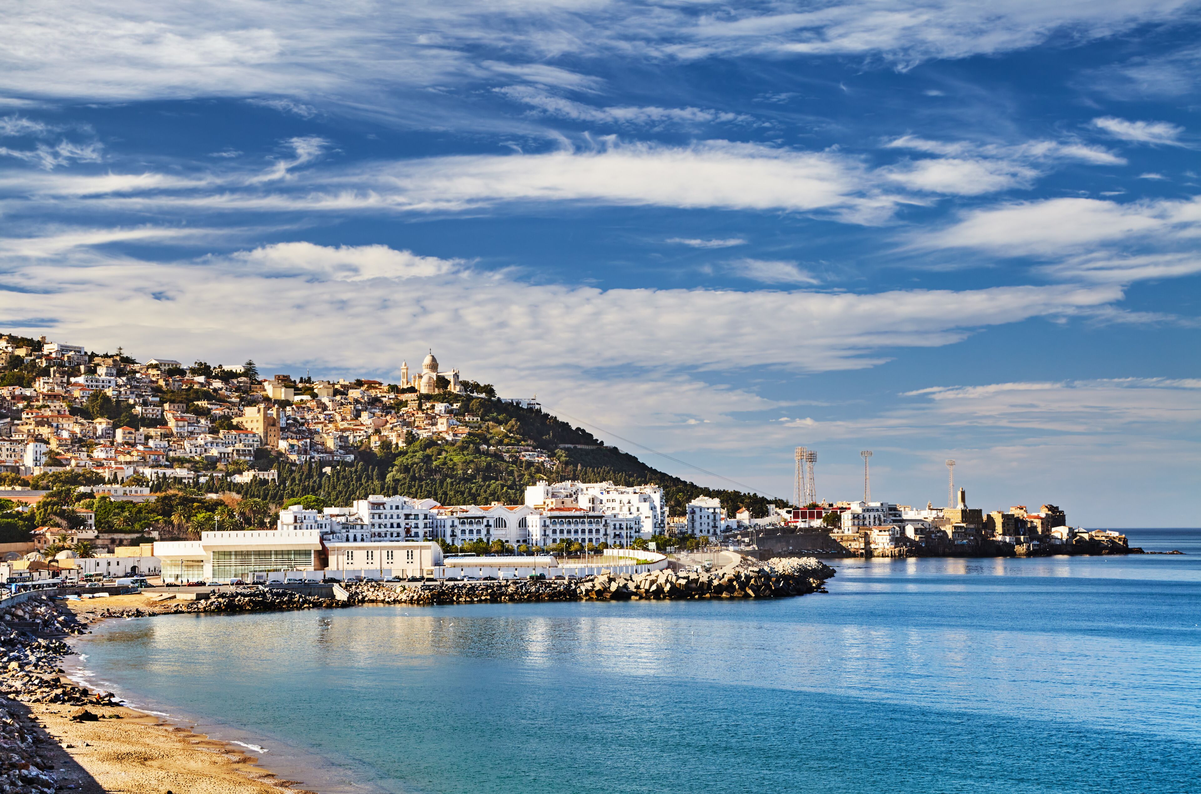 Uma vila costeira com edifícios numa encosta, uma praia e um céu azul límpido.