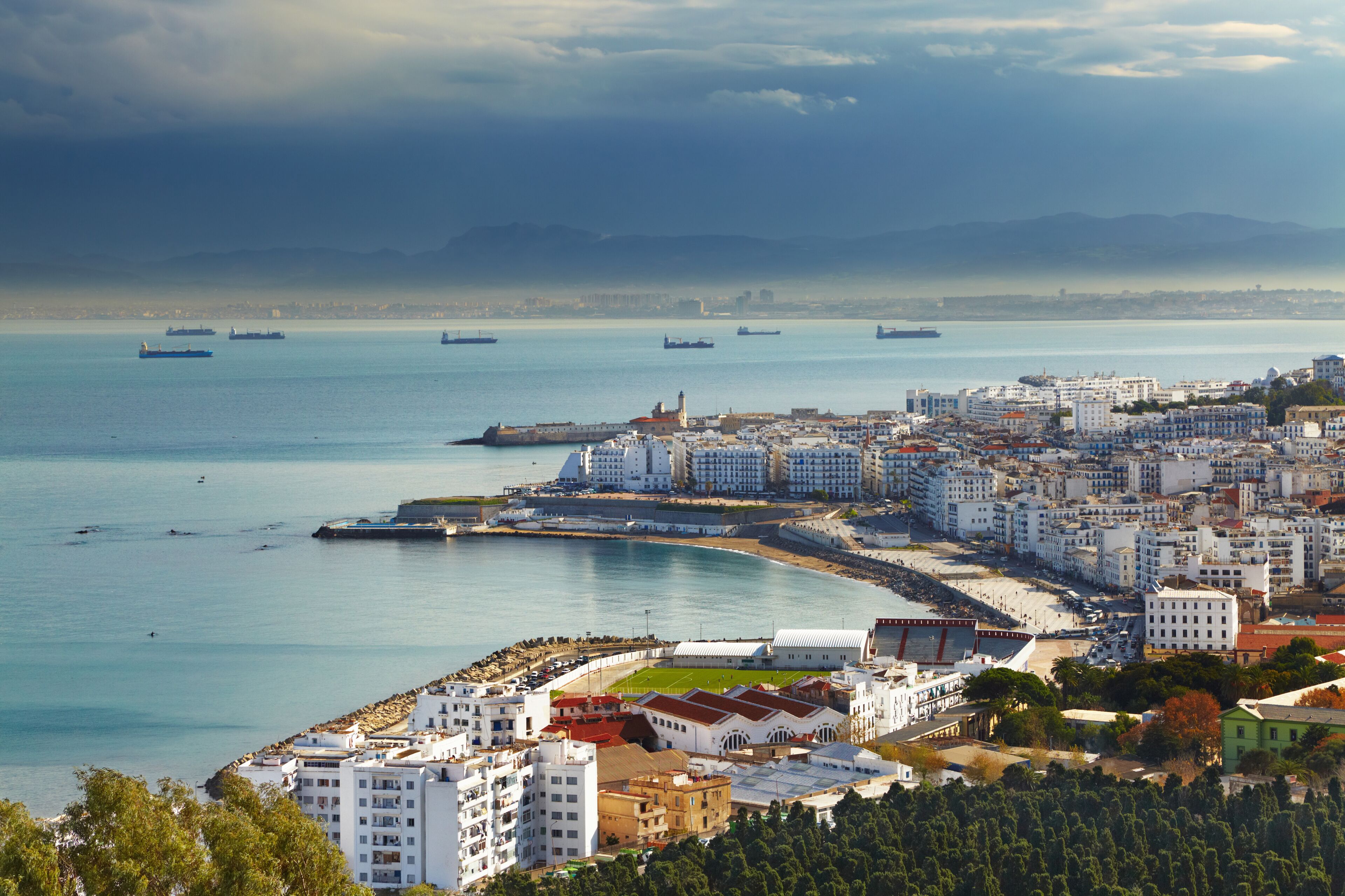 Uma cidade costeira com um porto, navios e um estádio.