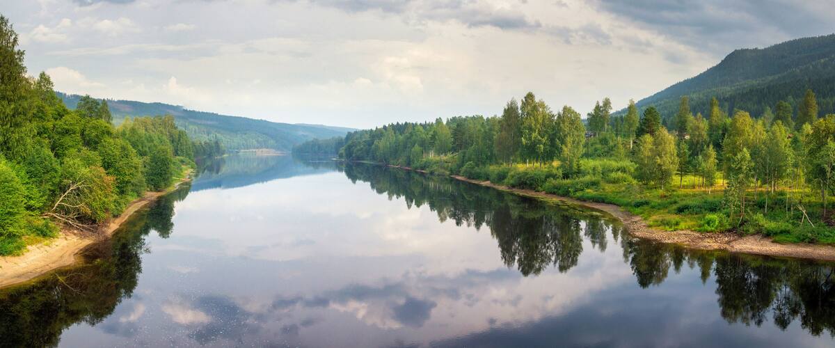 A calm river reflecting the surrounding greenery and a cloudy sky.