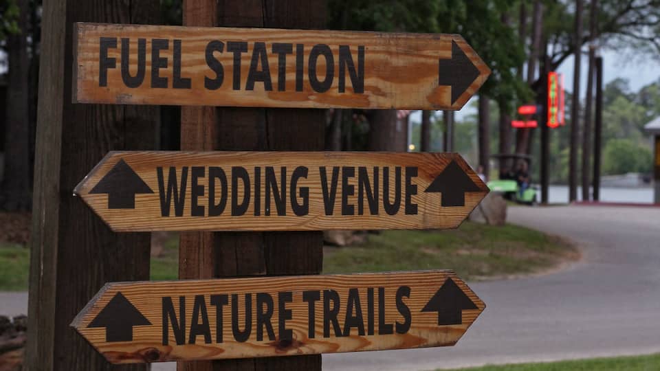 A wooden signpost with directional arrows pointing to various amenities such as cabins, swimming, restrooms, showers, fuel station, wedding venue, nature trails, playground, and glamping.