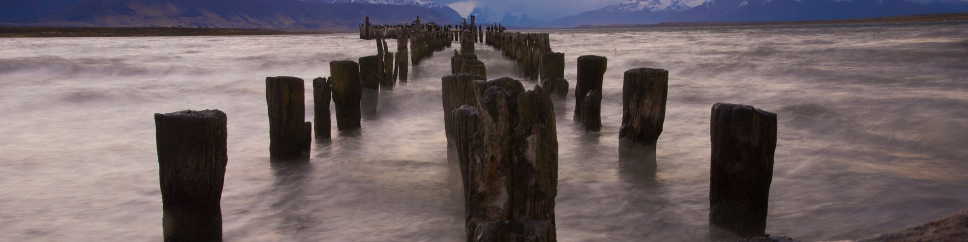 Un muelle de madera que se adentra en un lago tranquilo con montañas al fondo.