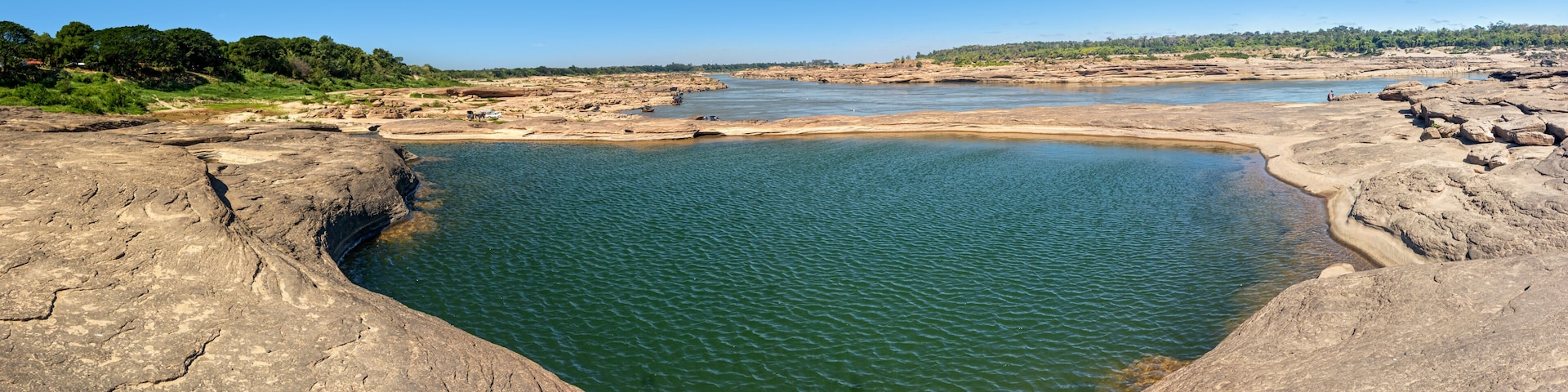 Una poza natural de roca rodeada de terreno pedregoso y vegetación distante.