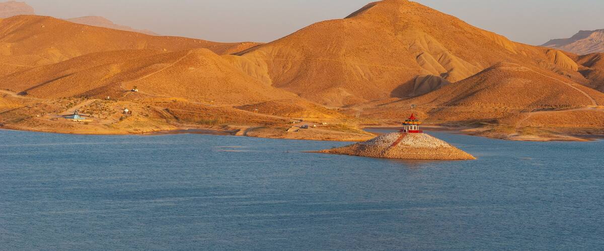 A small island with a lighthouse in the middle of a calm body of water, surrounded by a mountainous landscape.
