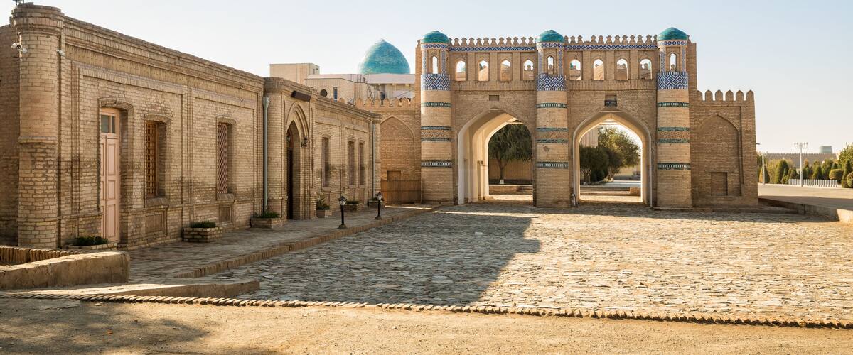 A historic stone archway with intricate patterns, leading to a courtyard with a dome in the background.